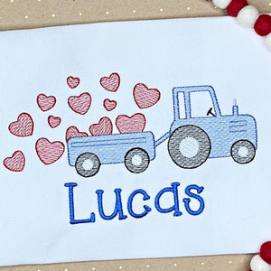 K&ouml;nnte beinhalten: Wei&szlig;es T-Shirt mit einem blauen Traktor, der einen Anh&auml;nger voller roter Herzen zieht. Der Name "Lucas" ist in Blau gestickt. Ein festliches Design f&uuml;r Valentinstag oder einen Kindergeburtstag.