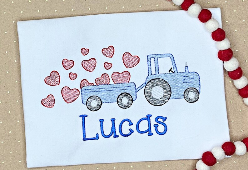 K&ouml;nnte beinhalten: Wei&szlig;es T-Shirt mit einem blauen Traktor, der einen Anh&auml;nger voller roter Herzen zieht. Der Name "Lucas" ist in Blau gestickt. Ein festliches Design f&uuml;r Valentinstag oder einen Kindergeburtstag.