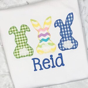 May include: White t-shirt featuring three appliqued Easter bunny designs in green, rainbow, and blue gingham patterns. The name "Reid" is embroidered in blue script below the bunnies. The bunnies have white lace accents.