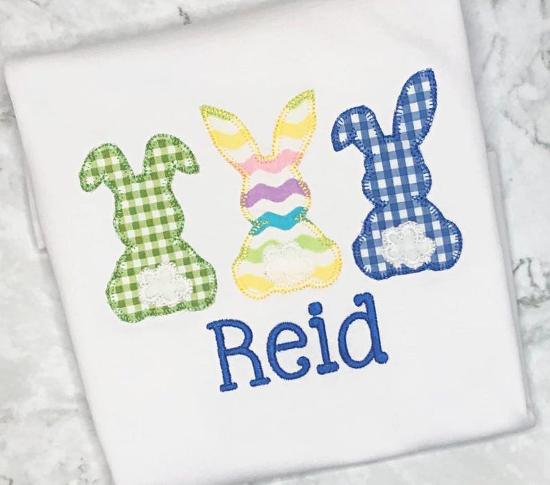 May include: White t-shirt featuring three appliqued Easter bunny designs in green, rainbow, and blue gingham patterns. The name "Reid" is embroidered in blue script below the bunnies. The bunnies have white lace accents.