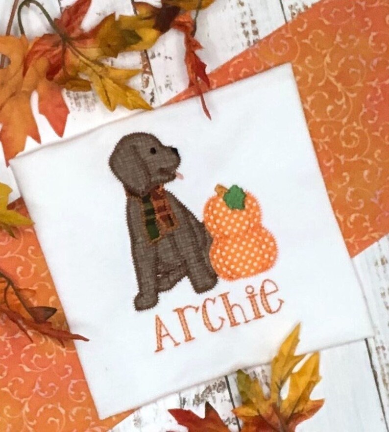 May include: White t-shirt featuring an embroidered brown dog wearing a scarf, with an orange polka-dot pumpkin. The name "Archie" is embroidered below the dog and pumpkin. Autumn leaves and orange fabric surround the shirt.