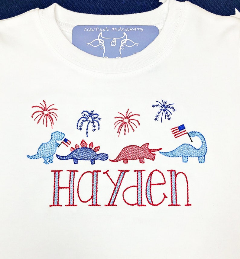 Puede incluir: Camiseta blanca con dinosaurios, fuegos artificiales y una bandera estadounidense bordados en rojo, blanco y azul. El nombre "Hayden" est&aacute; bordado debajo de los dinosaurios.