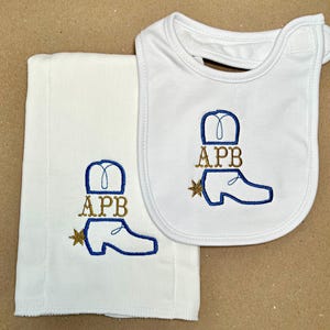 May include: A white burp cloth and bib with blue and gold embroidery of a cowboy boot with a star and the letters APB.