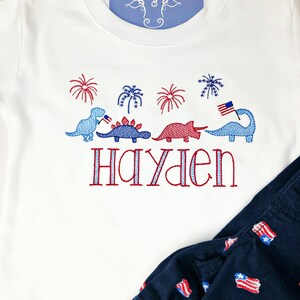Puede incluir: Camiseta blanca de manga corta con dinosaurios, fuegos artificiales y el nombre "Hayden" bordados en rojo, blanco y azul.