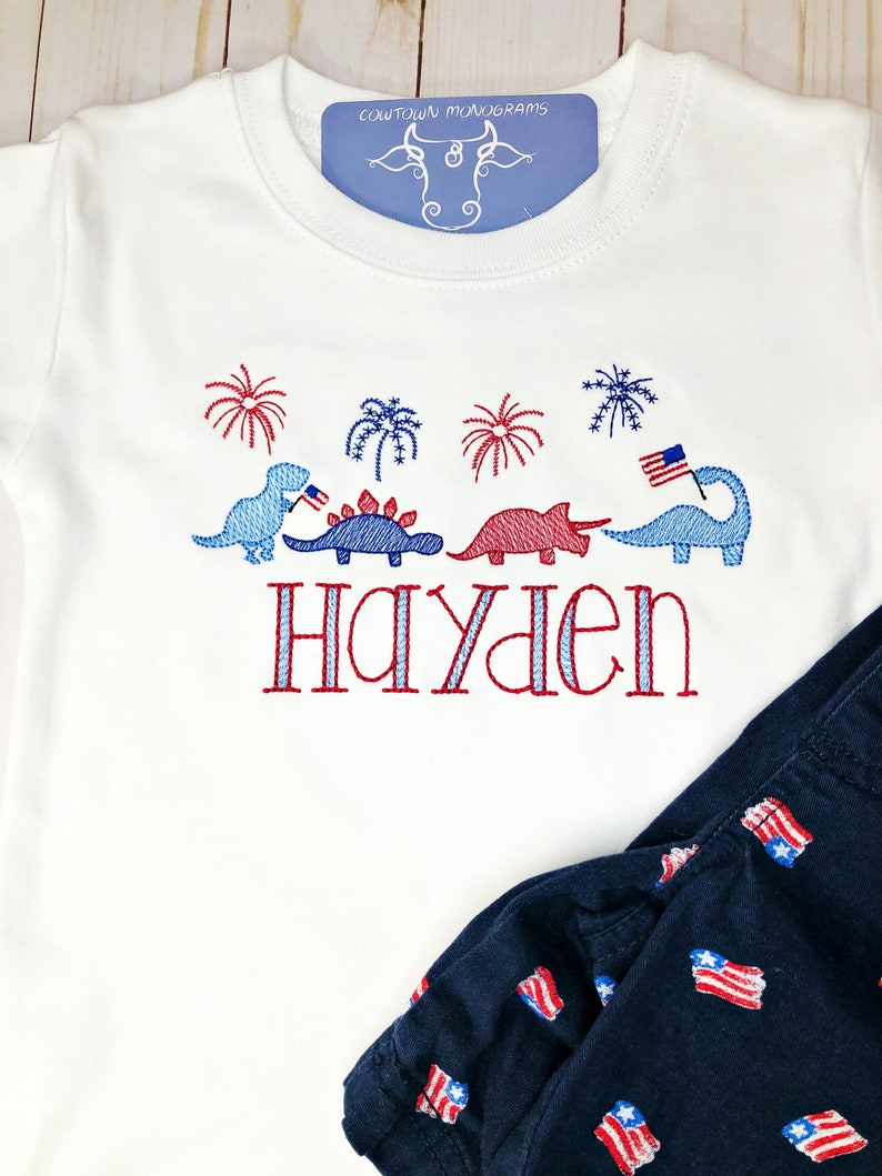 Puede incluir: Camiseta blanca de manga corta con dinosaurios, fuegos artificiales y el nombre "Hayden" bordados en rojo, blanco y azul.