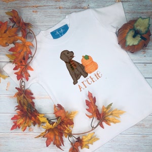 May include: White t-shirt featuring an embroidered brown dog wearing a plaid scarf, next to an orange pumpkin with a polka dot pattern. The name "Archie" is embroidered in orange below the dog and pumpkin. The shirt is decorated with fall leaves.
