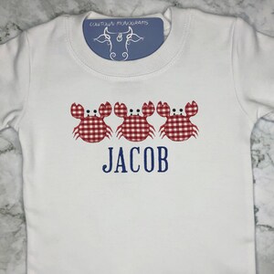 K&ouml;nnte beinhalten: Wei&szlig;er Baby-Strampler mit drei roten und wei&szlig;en Gingham-Krabben und dem Namen "JACOB" in Blau gestickt.