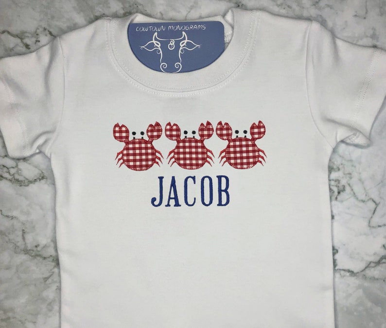 K&ouml;nnte beinhalten: Wei&szlig;er Baby-Strampler mit drei roten und wei&szlig;en Gingham-Krabben und dem Namen "JACOB" in Blau gestickt.