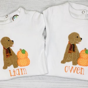 May include: Two white baby shirts with matching embroidered designs. Each shirt features a brown dog wearing a scarf next to an orange pumpkin. One shirt says "Liam" and the other says "Owen" in orange thread.