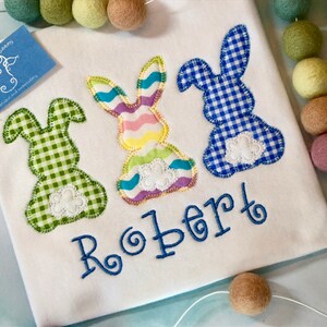 May include: White shirt with three appliqued Easter bunnies in green, pastel, and blue gingham patterns. The name "Robert" is embroidered in blue script below the bunnies. A small blue tag with a cow logo is in the upper left corner.