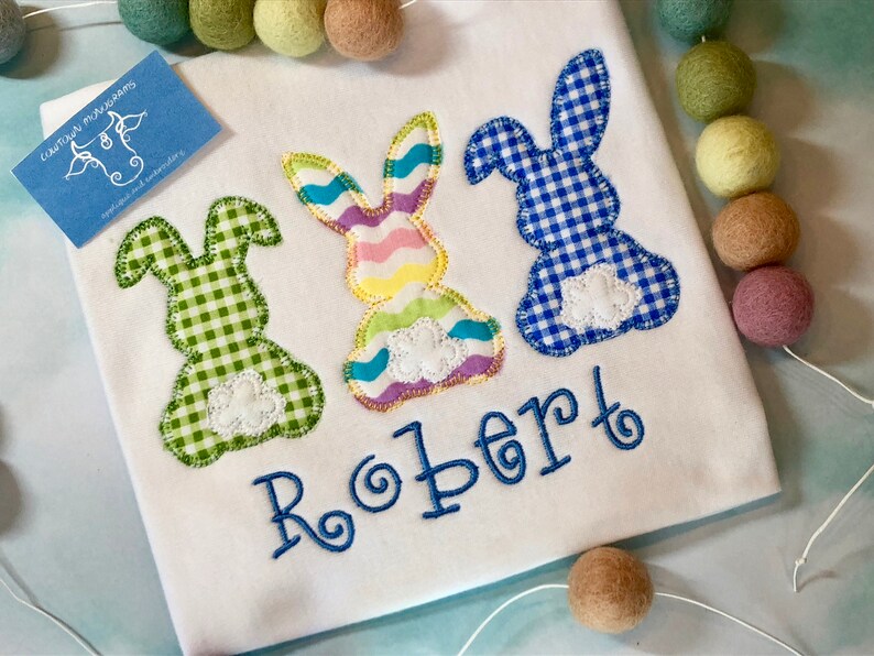 May include: White shirt with three appliqued Easter bunnies in green, pastel, and blue gingham patterns. The name "Robert" is embroidered in blue script below the bunnies. A small blue tag with a cow logo is in the upper left corner.