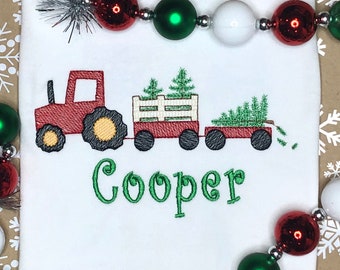 May include: Red toy tractor pulling a wagon with two green Christmas trees. The wagon has a white wooden fence. The name "Cooper" is embroidered in green below the tractor.