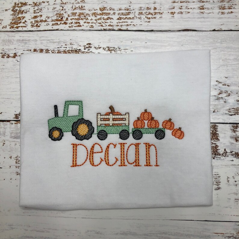 May include: White t-shirt with an embroidered design of a green tractor pulling two wagons filled with pumpkins. The name "Declan" is embroidered in orange below the tractor.