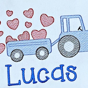 K&ouml;nnte beinhalten: Gesticktes Design eines blauen Traktors, der einen Wagen mit roten Herzen zieht. Der Name "Lucas" ist darunter in blauer Schrift gestickt. Das Design befindet sich auf wei&szlig;em Hintergrund.