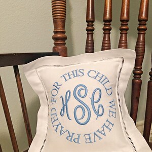 Puede incluir: Coj&iacute;n cuadrado blanco con texto e iniciales bordadas en azul. El texto dice "FOR THIS CHILD WE HAVE PRAYED". El coj&iacute;n est&aacute; sobre una silla de madera con un coj&iacute;n estampado. El bordado presenta las iniciales "HSE".