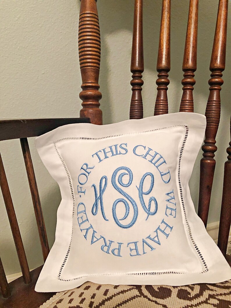 Puede incluir: Coj&iacute;n cuadrado blanco con texto e iniciales bordadas en azul. El texto dice "FOR THIS CHILD WE HAVE PRAYED". El coj&iacute;n est&aacute; sobre una silla de madera con un coj&iacute;n estampado. El bordado presenta las iniciales "HSE".