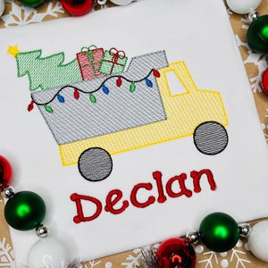 May include: White t-shirt with an embroidered yellow truck design. The truck carries a green Christmas tree, wrapped gifts, and fairy lights. The name "Declan" is embroidered in red. Festive holiday clothing.