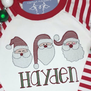 May include: White long-sleeved shirt with red and white striped sleeves. The shirt features three embroidered Santa faces and the name "Hayden" in green letters.