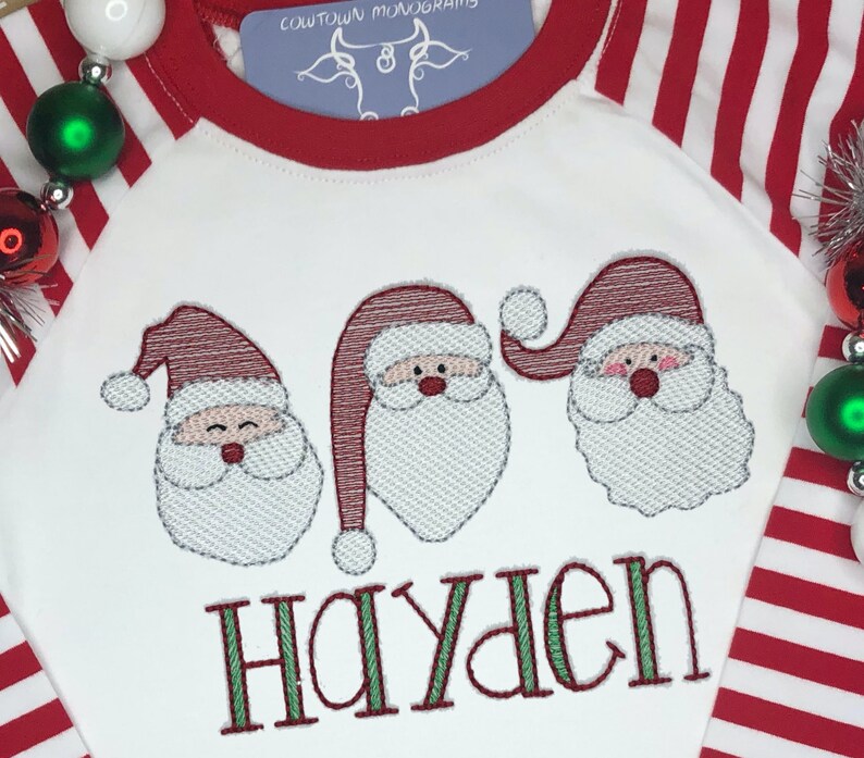 May include: White long-sleeved shirt with red and white striped sleeves. The shirt features three embroidered Santa faces and the name "Hayden" in green letters.