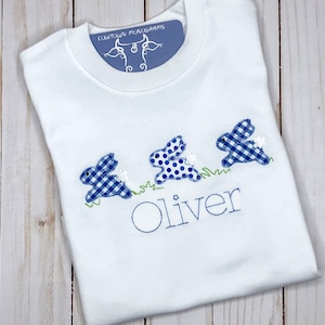 Camisa-body o babero personalizado del conejito de Pascua para niños, bordado con el trío de conejitos azules saltando por la hierba.