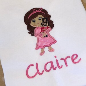 May include: Embroidered design of a girl in a pink dress holding a toy soldier, with the name "Claire" embroidered below.
