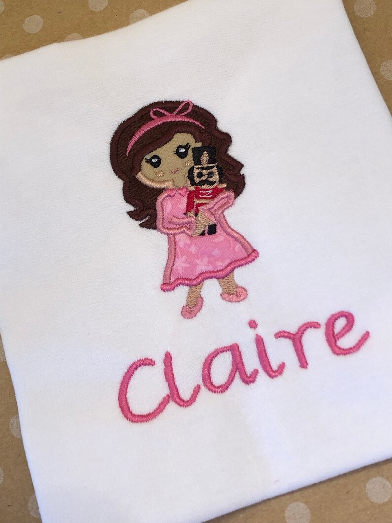 May include: Embroidered design of a girl in a pink dress holding a toy soldier, with the name "Claire" embroidered below.
