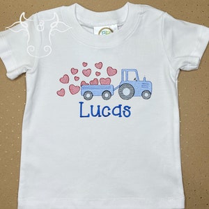 K&ouml;nnte beinhalten: Wei&szlig;es T-Shirt mit einem gestickten Design eines blauen Traktors, der einen Anh&auml;nger mit roten Herzen zieht. Der Name "Lucas" ist darunter blau gestickt. Das T-Shirt hat kurze &Auml;rmel und einen Rundhalsausschnitt.