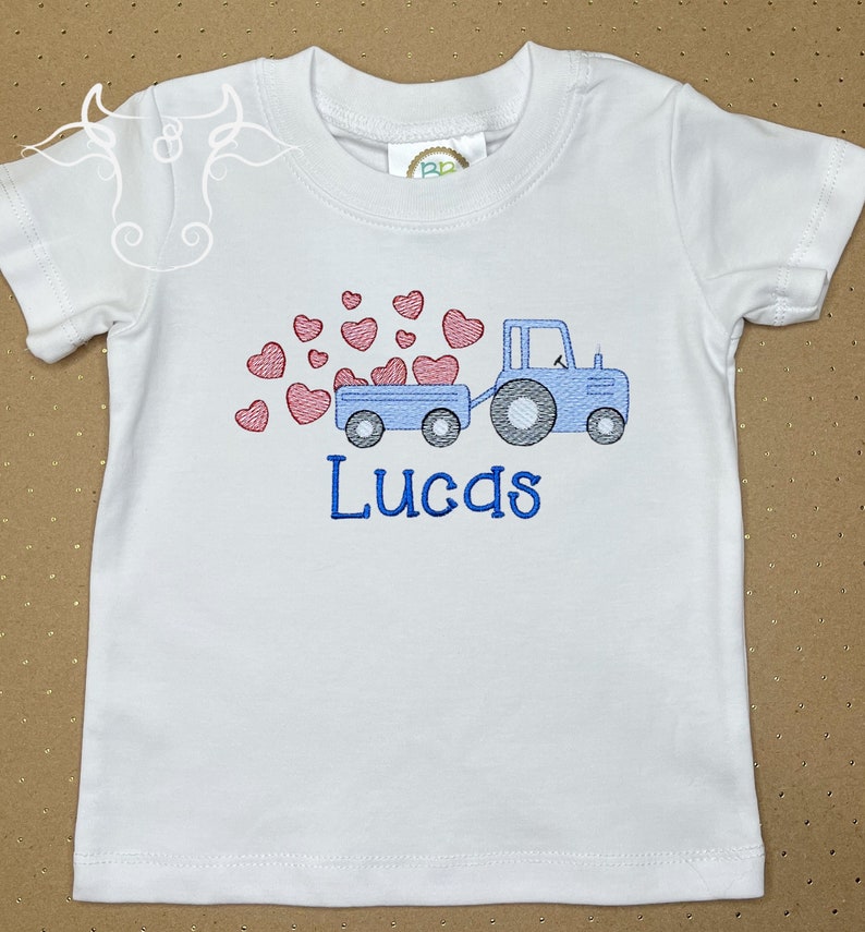 K&ouml;nnte beinhalten: Wei&szlig;es T-Shirt mit einem gestickten Design eines blauen Traktors, der einen Anh&auml;nger mit roten Herzen zieht. Der Name "Lucas" ist darunter blau gestickt. Das T-Shirt hat kurze &Auml;rmel und einen Rundhalsausschnitt.
