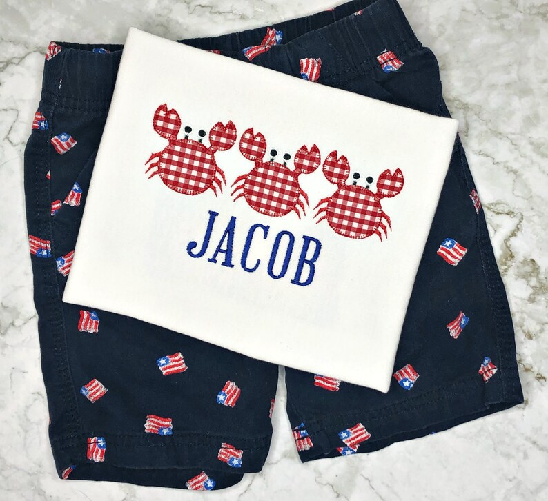 K&ouml;nnte beinhalten: Ein wei&szlig;es T-Shirt mit drei roten und wei&szlig;en Gingham-Krabben und dem Namen "JACOB" in Blau gestickt. Das Shirt wird mit dunkelblauen Shorts mit einem rot-wei&szlig;-blauen amerikanischen Flaggenmuster kombiniert.