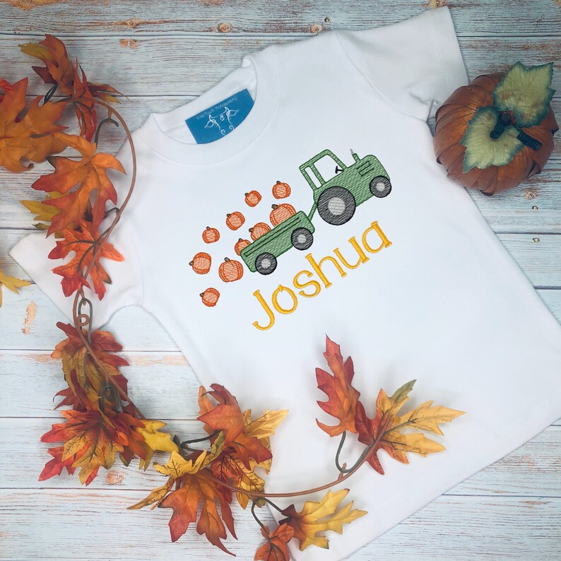 May include: A white t-shirt with a green tractor pulling a wagon full of orange pumpkins. The name "Joshua" is embroidered in yellow on the shirt.