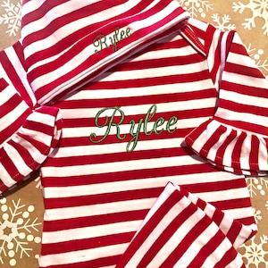 May include: Red and white striped baby outfit with a matching hat. The outfit has long sleeves and pants. The hat and outfit are embroidered with the name "Rylee" in green thread.