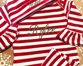May include: Red and white striped baby outfit with a matching hat. The outfit has long sleeves and pants. The hat and outfit are embroidered with the name "Rylee" in green thread.