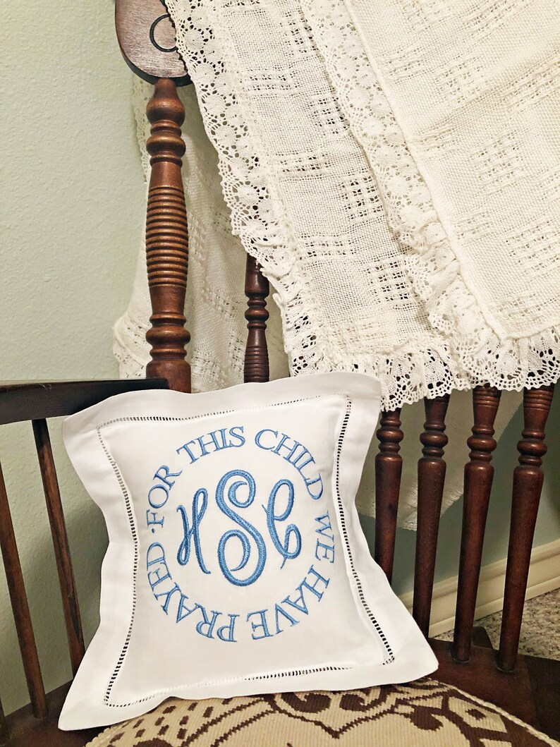 Puede incluir: Un coj&iacute;n cuadrado blanco con texto bordado en azul y un monograma, apoyado en una silla de madera. El texto dice "FOR THIS CHILD WE HAVE PRAYED". El coj&iacute;n tiene un borde cosido. Una manta de encaje color crema est&aacute; sobre la silla.