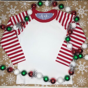 May include: White long-sleeved shirt with red and white striped sleeves. The shirt has a red collar and a small white label with the text "Couture Mushrooms" and a cow graphic. The shirt is surrounded by a garland of red, white and green Christmas baubles.