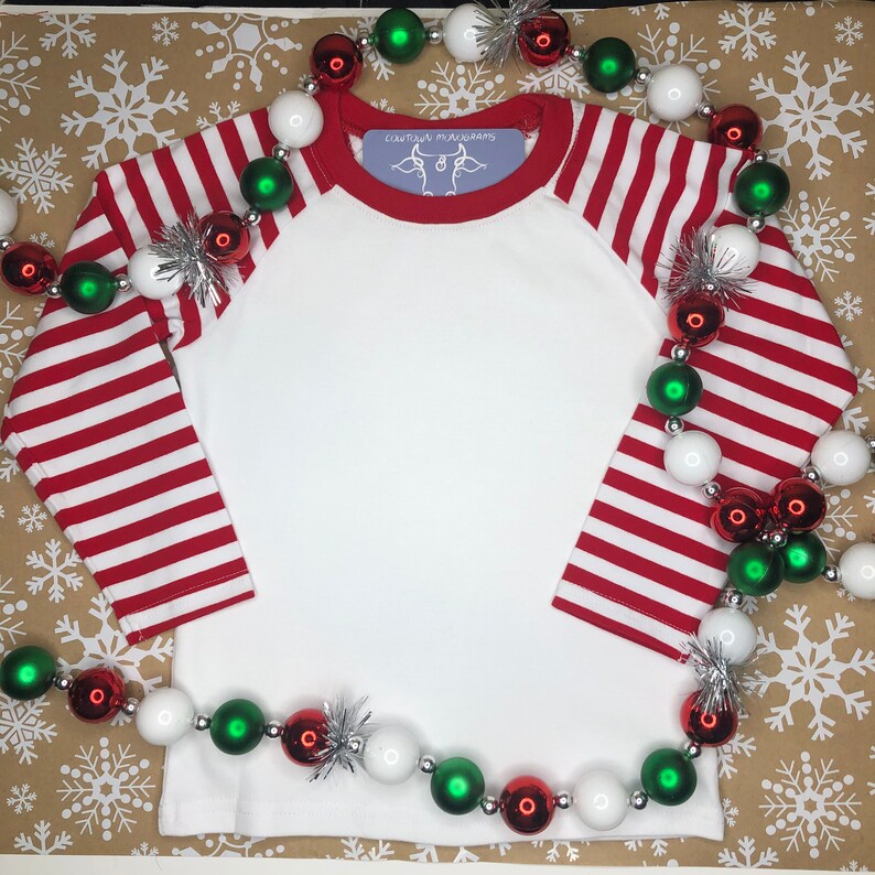 May include: White long-sleeved shirt with red and white striped sleeves. The shirt has a red collar and a small white label with the text "Couture Mushrooms" and a cow graphic. The shirt is surrounded by a garland of red, white and green Christmas baubles.