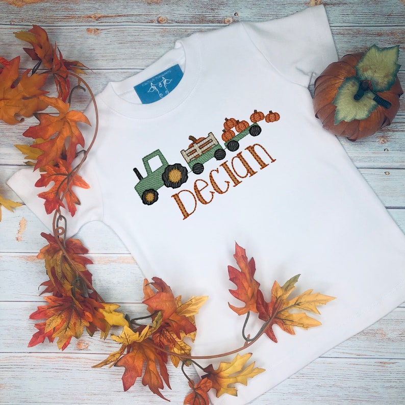 May include: White t-shirt with an embroidered design featuring a green tractor pulling a wagon filled with pumpkins. The name "Declan" is embroidered in orange. Autumn leaves and a pumpkin are in the background.