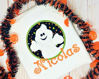 May include: White t-shirt with an embroidered ghost design inside a black circle with orange and yellow dots. The ghost has a smiling face. The name "Nicolas" is embroidered in orange script below the ghost. Halloween theme.