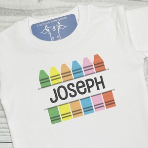 Könnte beinhalten: Weißes T-Shirt mit einem Design aus bunten Buntstiften und dem Namen "Joseph" in schwarzem Text.