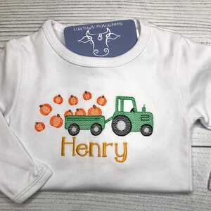May include: White long-sleeved shirt with a green tractor pulling a wagon full of orange pumpkins. The name "Henry" is embroidered in gold below the tractor.