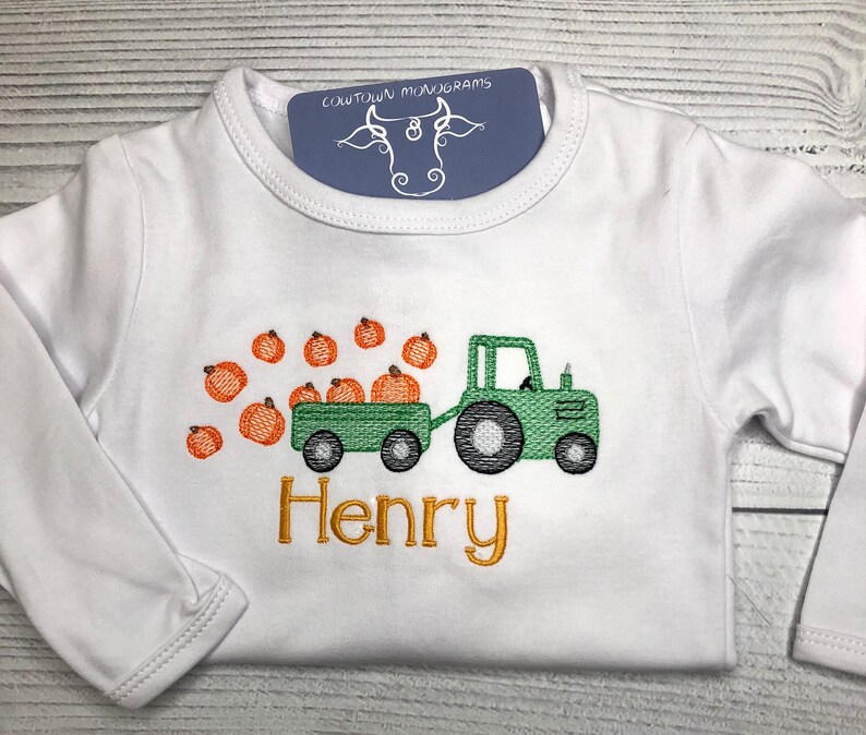 May include: White long-sleeved shirt with a green tractor pulling a wagon full of orange pumpkins. The name "Henry" is embroidered in gold below the tractor.