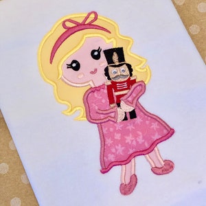 May include: Embroidered design of a blonde girl in a pink dress holding a toy nutcracker. The girl has a pink bow in her hair and the nutcracker has a red and black uniform.