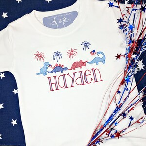 Puede incluir: Camiseta blanca con dinosaurios, fuegos artificiales y el nombre "Hayden" bordados en rojo, blanco y azul.