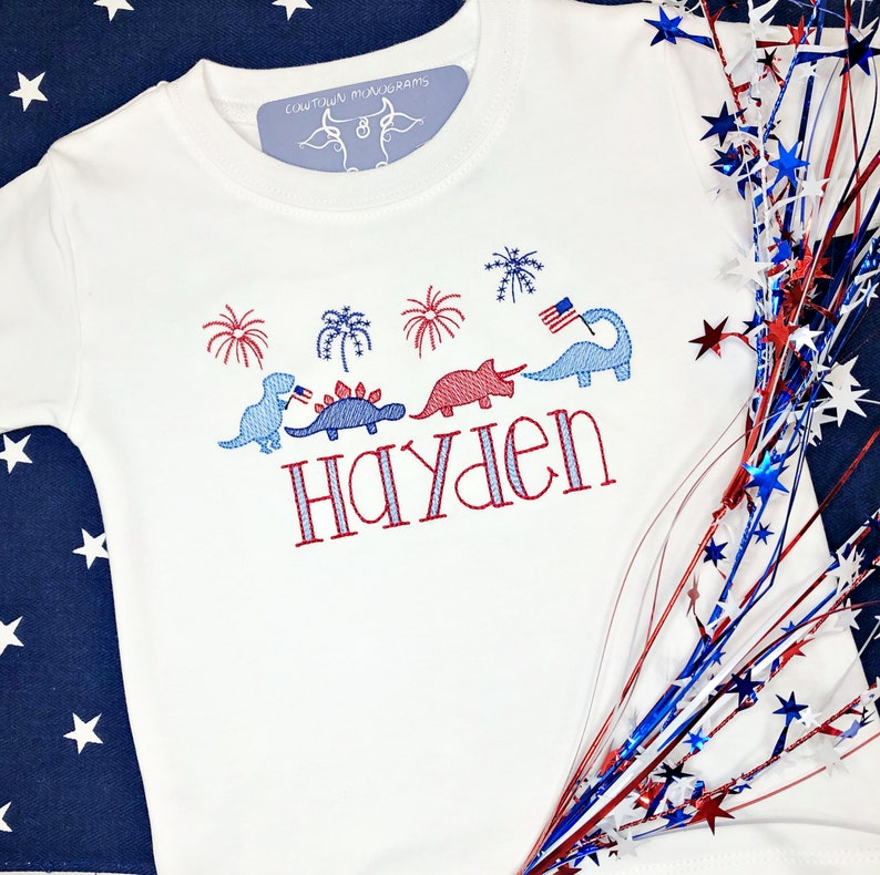 Puede incluir: Camiseta blanca con dinosaurios, fuegos artificiales y el nombre "Hayden" bordados en rojo, blanco y azul.