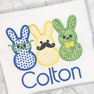 May include: White t-shirt featuring three Easter bunny designs. The bunnies are blue with glasses, yellow with a mustache, and green with a bow tie. The name "Colton" is embroidered in blue below the bunnies.