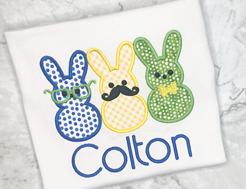 May include: White t-shirt featuring three Easter bunny designs. The bunnies are blue with glasses, yellow with a mustache, and green with a bow tie. The name "Colton" is embroidered in blue below the bunnies.