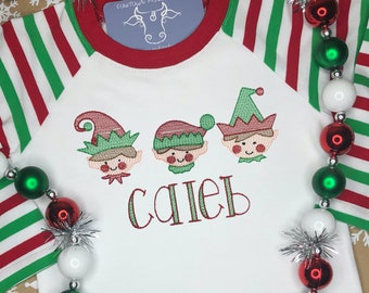 Puede incluir: Camiseta blanca de manga larga con mangas a rayas rojas y verdes. La camiseta presenta un diseño bordado de tres elfos con el nombre "Caleb" bordado debajo.