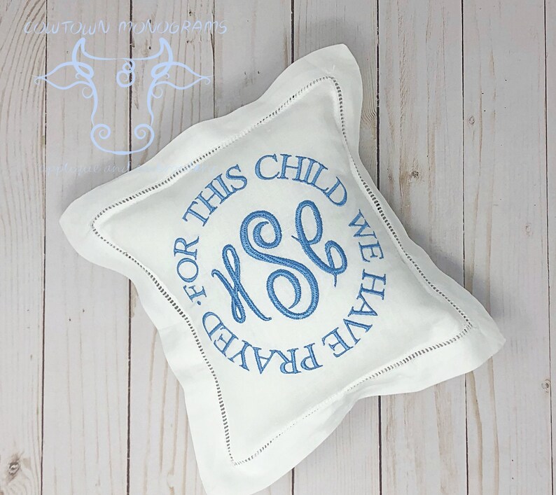 Puede incluir: Almohada cuadrada blanca con texto bordado azul y monograma. El texto dice "FOR THIS CHILD WE HAVE PRAYED". La almohada tiene un borde cosido decorativo y est&aacute; sobre una superficie de madera clara.
