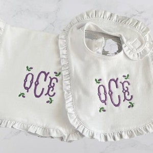 May include: A set of two white baby bibs with ruffled edges. Both bibs feature a purple embroidered monogram "OCE" with green leaves.