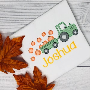 May include: A white t-shirt with a green tractor pulling a wagon full of orange pumpkins. The tractor has a black tire and the wagon has a black wheel. The text "Joshua" is embroidered in yellow on the shirt.
