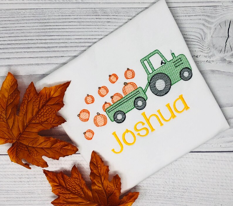 May include: A white t-shirt with a green tractor pulling a wagon full of orange pumpkins. The tractor has a black tire and the wagon has a black wheel. The text "Joshua" is embroidered in yellow on the shirt.
